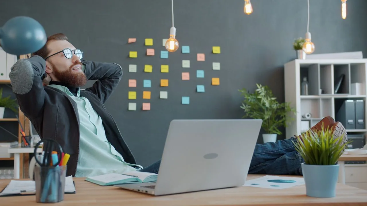 A content lead leaning back at their desk beside a laptop and a wall of sticky notes